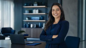Mulher adulta sorridente em ambiente de escritório moderno, sentada em uma cadeira com braços cruzados, usando blazer azul e gestos de confiança diante de um computador na mesa ao fundo com benefícios corporativos no contrato intermitente.