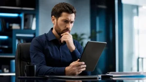Homem trabalhando em escritório moderno, sentado à mesa com computador, digitando e pensando sobre a multa por não cadastrar trabalhador intermitente no eSocial, com a mão no queixo, ambiente corporativo com luz natural ao fundo.