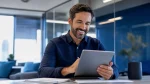 Homem sorridente em escritório corporativo, usando um tablet e trabalhando em frente ao computador lendo sobre salário do trabalhador intermitente, em um ambiente moderno com vidros e mesas.
