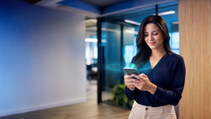 Mulher em ambiente corporativo usando smartphone, com ícone de verificação na tela, sugerindo controle de ponto com geolocalização e registro de presença em tempo real.