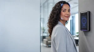 Mulher com jaleco sorrindo em um ambiente de escritório, ao lado de um terminal de controle de ponto com reconhecimento facial instalado na parede, com luzes de leitura no dispositivo.