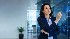 Mulher executiva em escritório moderno, apontando para um painel com gráficos e linhas digitais, sugerindo diagnóstico de eficiência em processos e desempenho.