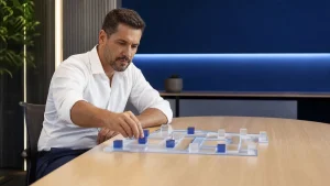 Homem em escritório analisa um tabuleiro com peças e blocos azuis e brancos sobre a mesa, enquanto faz um cálculo cuidadoso, representando um diagnóstico de eficiência.