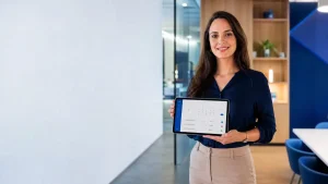 Mulher em ambiente corporativo apresenta um tablet com interface de controle de atividades e horários, sugerindo um sistema de ponto eletrônico para empresas no escritório.